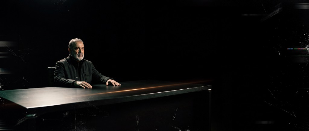 A man seated behind a long dark desk under a single spotlight, an amber light streak across the desk.