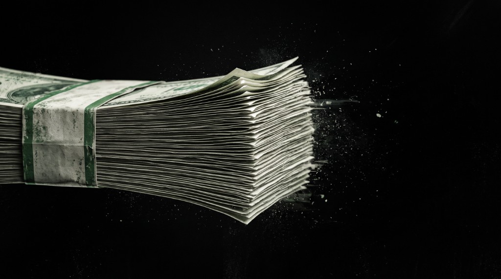 A banded stack of cash dissolving into particles against black.